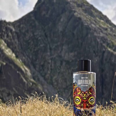 Sticlă de aftershave Marmara Barber Columbia așezată pe iarbă uscată, cu un munte stâncos în fundal.